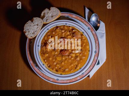 TYPISCH SPANISCHE FABADA AUF EINEM TELLER, MIT ZWEI BROTSTÜCKEN UND EINEM LÖFFEL AUF EINER SERVIETTE. ALLES IST ÜBER EINEM HOLZTISCH Stockfoto