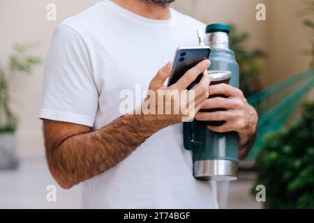 Mann mit Kürbis und Thermoskanne, der ein Handy in der Hand hält. Traditionelles südamerikanisches Heißgetränk. Stockfoto
