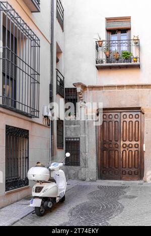 Weißer Roller parkt in einem schmalen Innenhof des Wohnviertels von Toledo, Spanien. Gebäudefassaden mit traditionellen Balkonen und Fenstern. Stockfoto