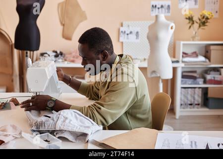 Seitenansicht eines jungen afroamerikanischen Handwerkers, der an einer elektrischen Nähmaschine im Studio langsamer Mode sitzt und neue Kleidung kreiert Stockfoto
