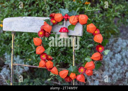 Kranz aus Physalis, Strohblumen und Efeublättern, verziert mit einer Holzfliege, die auf einem Gartenstuhl hängt Stockfoto