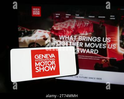 Person mit Smartphone und Logo der Schweizer Automobilausstellung Genf International Autosalon vor der Website. Schwerpunkt auf dem Display des Telefons. Stockfoto