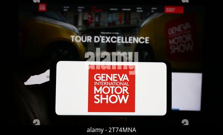 Person, die ein Mobiltelefon mit dem Logo der Schweizer Autosalon Genf vor der Business-Webseite hält. Schwerpunkt auf dem Display des Telefons. Stockfoto