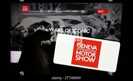 Person mit Handy mit Logo der Schweizer Autosalon Genf International Autosalon vor der Business-Webseite. Schwerpunkt auf dem Display des Telefons. Stockfoto