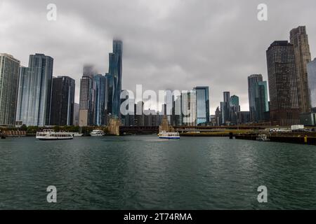 Das St. Regis Chicago erhebt sich über der Skyline von Chicago, Illinois, USA, sein Turmspitze ist in niedrige Wolken gehüllt. Der 2020 fertiggestellte 365-Meter-(1.198-Fuß)- Wolkenkratzer der Architektin Jeanne Gang gilt als das höchste Gebäude der Welt, das von einer Frau entworfen wurde. Zwei große weiße Ausflugsboote kreuzen die Mündung des Chicago River im Vordergrund, kurz vor der Brücke, an der der Fluss auf den offenen See trifft. Stockfoto