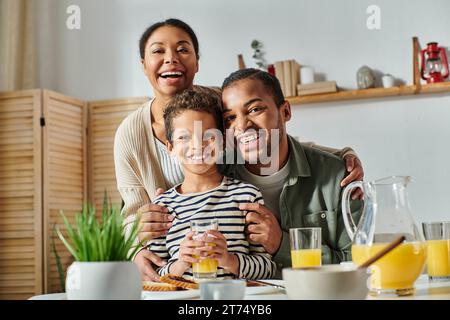 Fröhliche afroamerikanische Familie umarmt sich warm und lächelt fröhlich vor der Kamera am Frühstückstisch Stockfoto