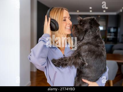 Glückliche junge Frau, die Kopfhörer trägt und zu Hause einen Schnauzer umarmt Stockfoto