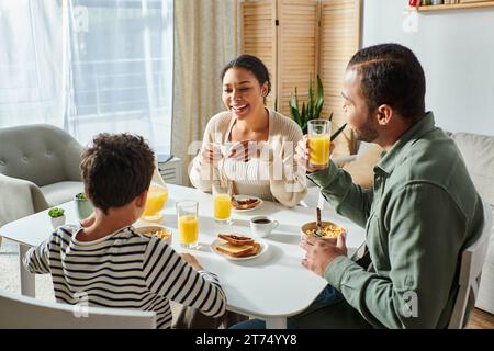Fröhliche, moderne afroamerikanische Familie, die zusammen frühstückt und sich anlächelt Stockfoto