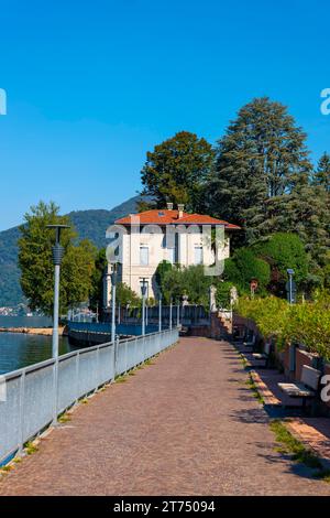 Gehweg mit Lampen und Geländer und ein Haus am Ufer am Luganer See mit Bergen und Bäumen an einem sonnigen Sommertag in Porto Ceresio, Lombardei Stockfoto