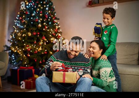 Glücklicher afroamerikanischer Mann, der ein Geschenk auspackt, während seine Frau und sein Sohn ihn anlächeln, Weihnachten Stockfoto