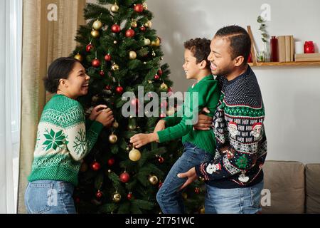 Fröhliche afroamerikanische Mutter, die ihren Mann ansieht und ihrem Sohn dabei hilft, den Weihnachtsbaum zu schmücken Stockfoto
