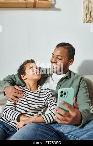 Vertikale Aufnahme eines fröhlichen afroamerikanischen Jungen, der seinen Vater fröhlich anlächelt, während er Selfies macht Stockfoto