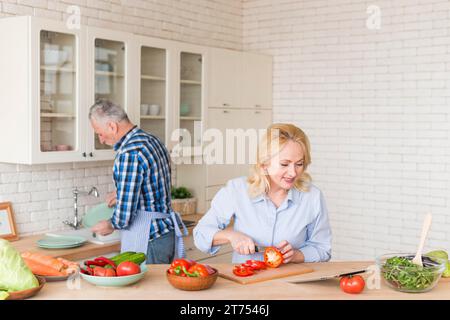Lächelnde Seniorin schneidet rote Glockenpfeffer mit einem Messer aussehenden digitalen Tablet, sein Mann wäscht Geschirr Küchenspüle Stockfoto