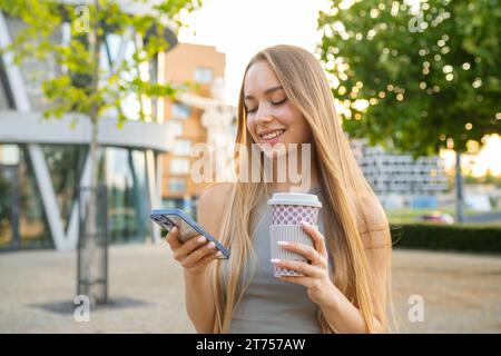 Die attraktive blonde Frau benutzt ihr Smartphone und trinkt Kaffee auf der Straße. Stockfoto