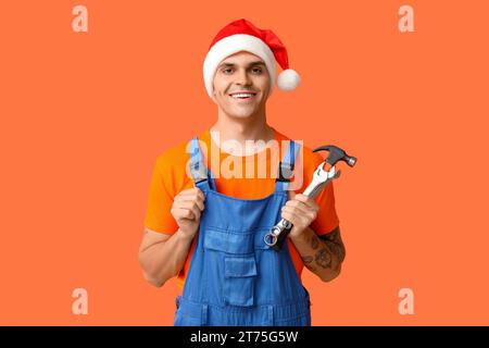 Männlicher Klempner im Weihnachtsmann-Hut mit Hammer und Schraubenschlüsseln auf orangem Hintergrund Stockfoto