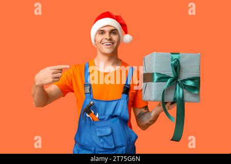 Männlicher Klempner im Weihnachtsmann-Hut mit Werkzeugen, die auf eine Geschenkbox auf orangem Hintergrund zeigen Stockfoto
