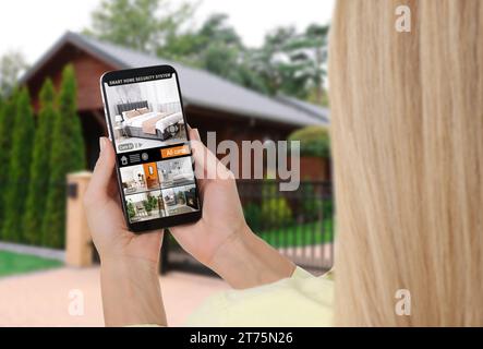 Frau, die ein intelligentes Heimsicherheitssystem auf dem Handy in der Nähe des Hauses im Freien benutzt. Gerät, das verschiedene Räume durch Kameras anzeigt Stockfoto