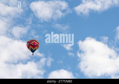 Ein isolierter, mehrfarbiger Heißluftballon schwimmt in der Luft mit weißen Wolken und einem strahlend blauen Himmel. Stockfoto