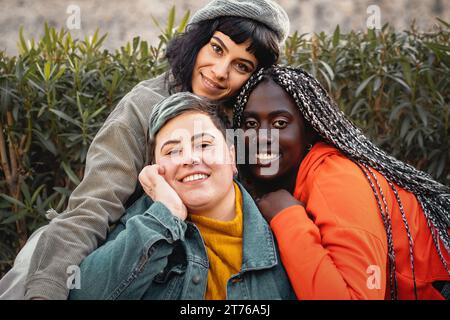 Drei enge Freunde teilen sich eine warme Umarmung und zeigen ihre einzigartigen Styles und ihr fröhliches Lächeln vor einer natürlichen Kulisse. Stockfoto