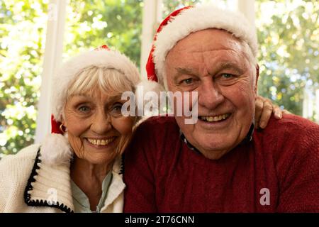 Glückliches weißes Seniorenpaar in santa Hats, das weihnachten Videoanruf hat und im sonnigen Zimmer lächelt Stockfoto