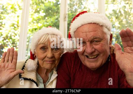 Glückliches kaukasisches Seniorenpaar in santa Hats, das weihnachten Videoanruf hat und im sonnigen Zimmer winkt Stockfoto