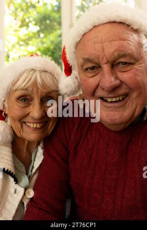 Glückliches weißes Seniorenpaar in santa Hats, das weihnachten Videoanruf hat und im sonnigen Zimmer lächelt Stockfoto