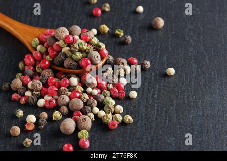 Farbige Paprika mischen sich in Holzlöffel und verstreut in der Nähe auf schwarzem Stein Hintergrund Nahaufnahme Stockfoto