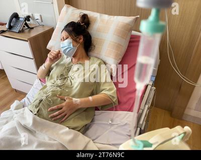 Schwangere Patientin in medizinischer Maske Husten auf einem Bett im Krankenhaus Stockfoto