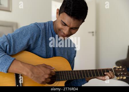 Glücklicher birassischer Mann, der zu Hause auf dem Bett sitzt und Gitarre spielt Stockfoto
