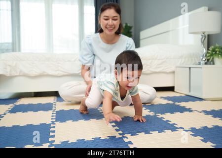 Glückliche Mutter, die ihr Baby auf der Spielmatte oder dem Boden im Schlafzimmer krabbelt Stockfoto