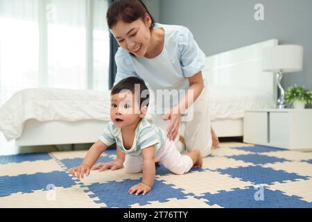 Glückliche Mutter, die ihr Baby auf der Spielmatte oder dem Boden im Schlafzimmer krabbelt Stockfoto