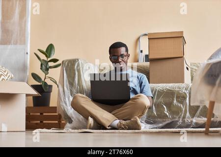 Junger seriöser Geschäftsmann mit Brille, der auf dem Boden sitzt und auf den Laptop-Bildschirm schaut, während er zu Hause Networking oder Online-Videos ansieht Stockfoto