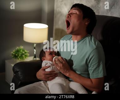 Verschlafener Vater gähnt und füttert das Baby nachts auf einem Sofa im Wohnzimmer mit einer Milchflasche Stockfoto