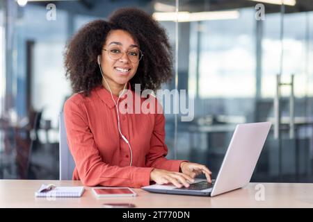 Porträt der jungen schönen erfolgreichen Frau am Arbeitsplatz im Büro, Geschäftsfrau im Kopfhörer lächelnd in die Kamera, zufrieden mit Leistung Ergebnisse Programmierer mit Laptop. Stockfoto