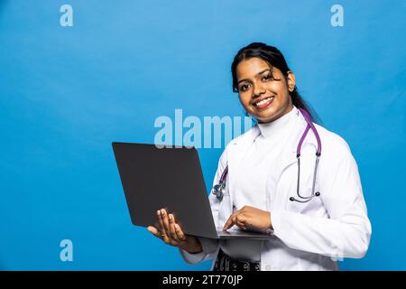 Fröhliche junge attraktive hinduistische Ärztin in Uniform arbeitet am Computer isoliert auf weißem Hintergrund. Soziale Distanz während covid-19-Pandem Stockfoto