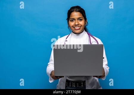 Fröhliche junge attraktive hinduistische Ärztin in Uniform arbeitet am Computer isoliert auf weißem Hintergrund. Soziale Distanz während covid-19-Pandem Stockfoto