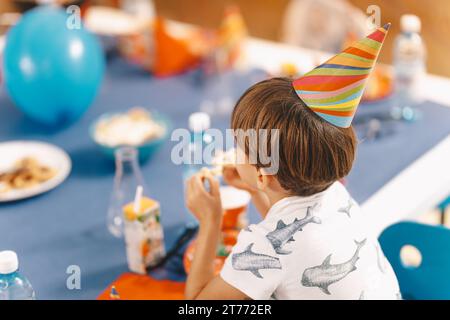 Kindergeburtstag. Kleiner Junge, der einen Geburtstagskegelmütze trägt. Anonymer Schuljunge sitzt am Tisch und isst Geburtstagskuchen Stockfoto