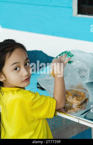 Ich nehme frittierte Speisen aus der Plastiktüte auf dem Tisch. Asiatischer Junge, der in die Kamera schaut. Stockfoto