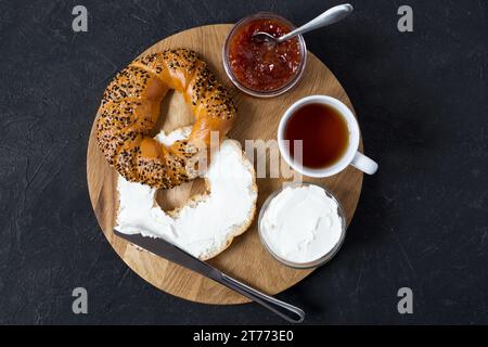 Frisch gebackener Bagel mit Frischkäse, Marmelade und einer Tasse Tee. Frühstück auf dem Holztablett auf dunklem Hintergrund. Draufsicht. Stockfoto