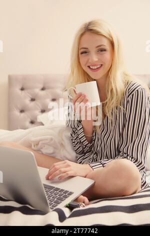 Lächelnde junge Frau sitzt auf Bett mit Laptop Stockfoto