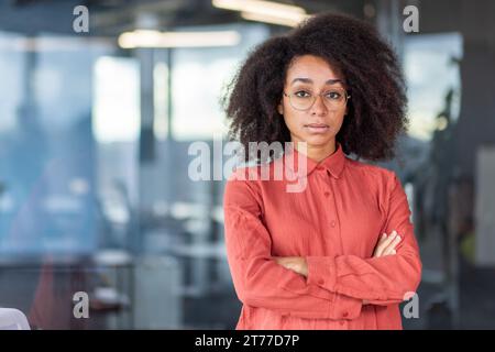 Porträt einer ernsthaften selbstbewussten Geschäftsfrau im Büro am Arbeitsplatz, einer Arbeiterin, die am Fenster steht und die Arme kreuzt und in die Kamera fokussiert. Stockfoto