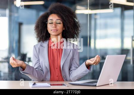 Ernsthaft denkende Frau, die sich in Lotusposition im Büro entspannt, Geschäftsfrau mit geschlossenen Augen am Schreibtisch mit Laptop sitzt. Stockfoto