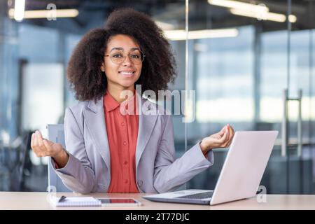 Porträt einer glücklichen Frau in Lotuspose, die im Büro am Arbeitsplatz meditiert, Geschäftsfrau lächelt und in die Kamera schaut, mit Laptop arbeitet. Stockfoto