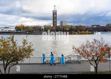 Am Tag der Eröffnung der Karnevalsveranstaltung am 11.11.23 spaziert eine Gruppe von Menschen in blauen Overalls am Ufer des Rheins entlang, dem alten Turm des f Stockfoto