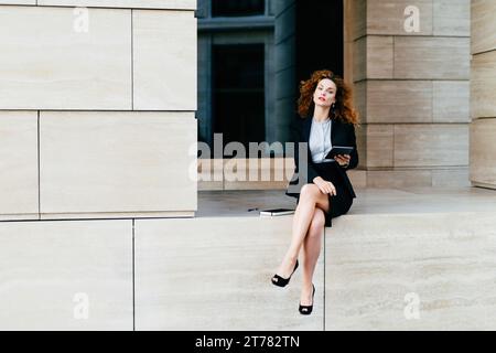 Elegante Geschäftsfrau im Anzug im Freien, mit einem Tablet in der Hand und einem Notizbuch neben ihr, verkörpert Professionalität Stockfoto