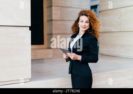 Selbstbewusste Geschäftsfrau mit lebhaftem lockigem Haar, die ein Tablet vor einem modernen Hintergrund hält Stockfoto