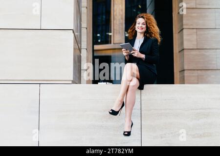 Fröhliche Geschäftsfrau mit lockigen Haaren, die ein Tablet benutzt, sitzt auf einem Vorsprung vor einem modernen Gebäude Stockfoto