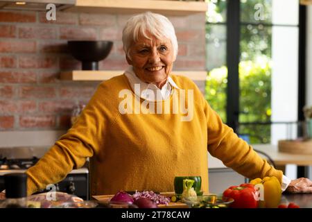 Glückliche kaukasische Seniorin, die mit frischem Gemüse auf der Theke in der sonnigen Küche steht Stockfoto