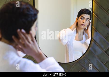 Glückliche birassische Frau, die Haare berührt und zu Hause im Badezimmerspiegel reflektiert Stockfoto