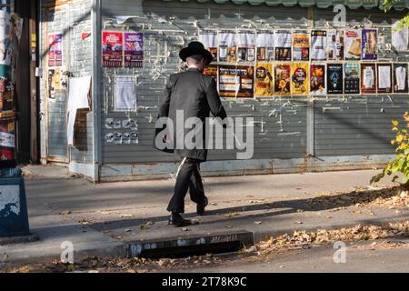 Ein chassidischer jüdischer Mann geht an einer Mauer von Ankündigungen und Anzeigen vorbei, die meist auf Jiddisch sind. Auf der Hooper Street in Williamsburg, Brooklyn, New York. Stockfoto